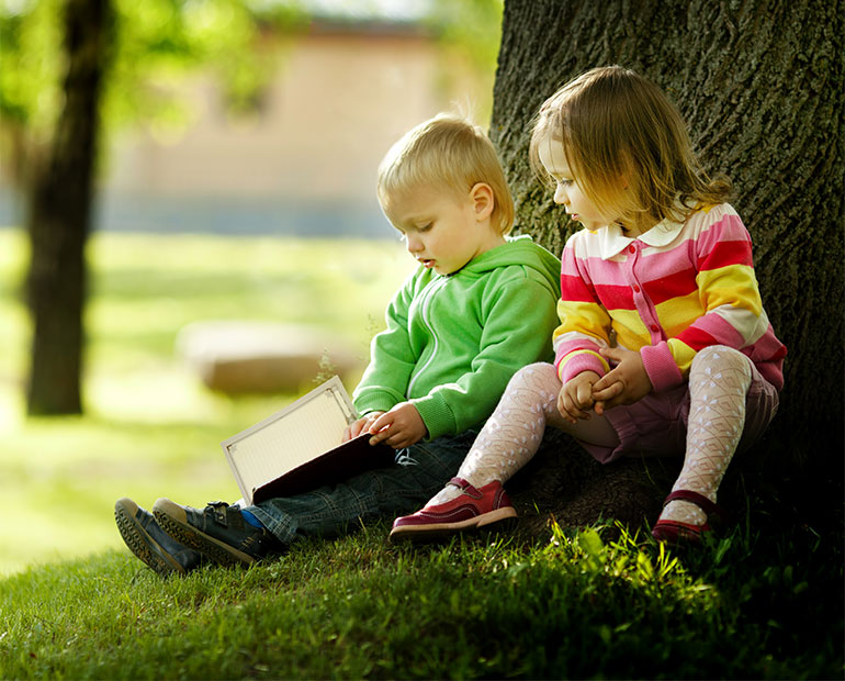 A toddler boy and a little girl reading by a tree. 